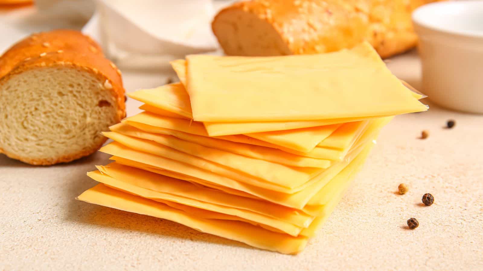 A stack of sliced cheese is placed on a light surface. Surrounding it are portions of sliced bread, a few peppercorns, and a small, white dish. The scene suggests preparation for a meal or snack.