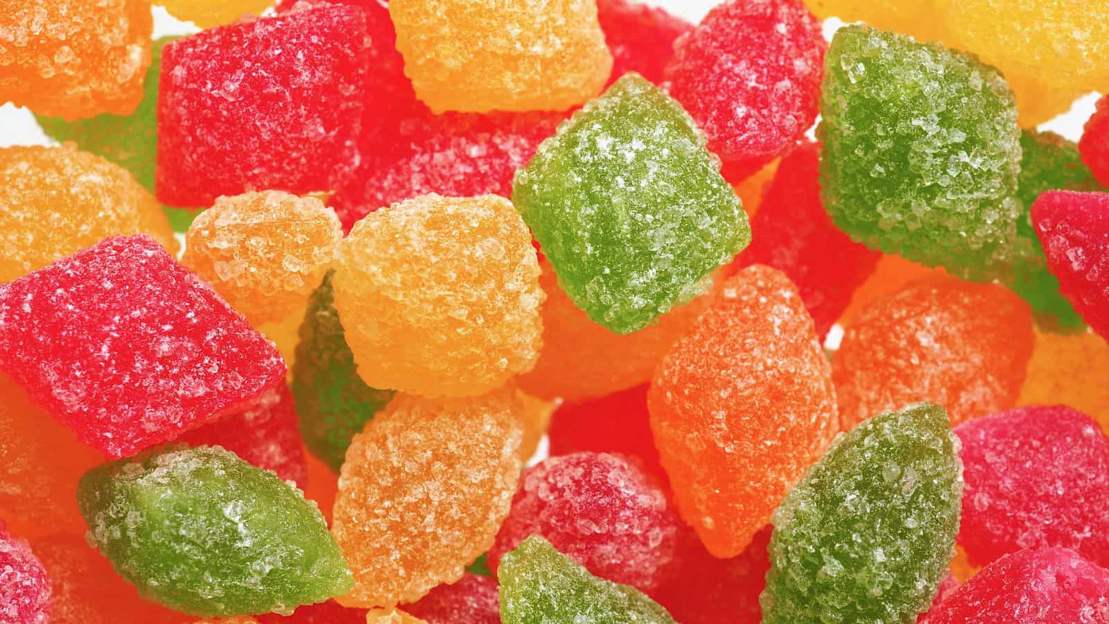 A close-up image of assorted colorful gummy candy pieces coated in sugar. The candies are in shades of red, green, orange, and yellow, with a granular texture from the sugar crystals.