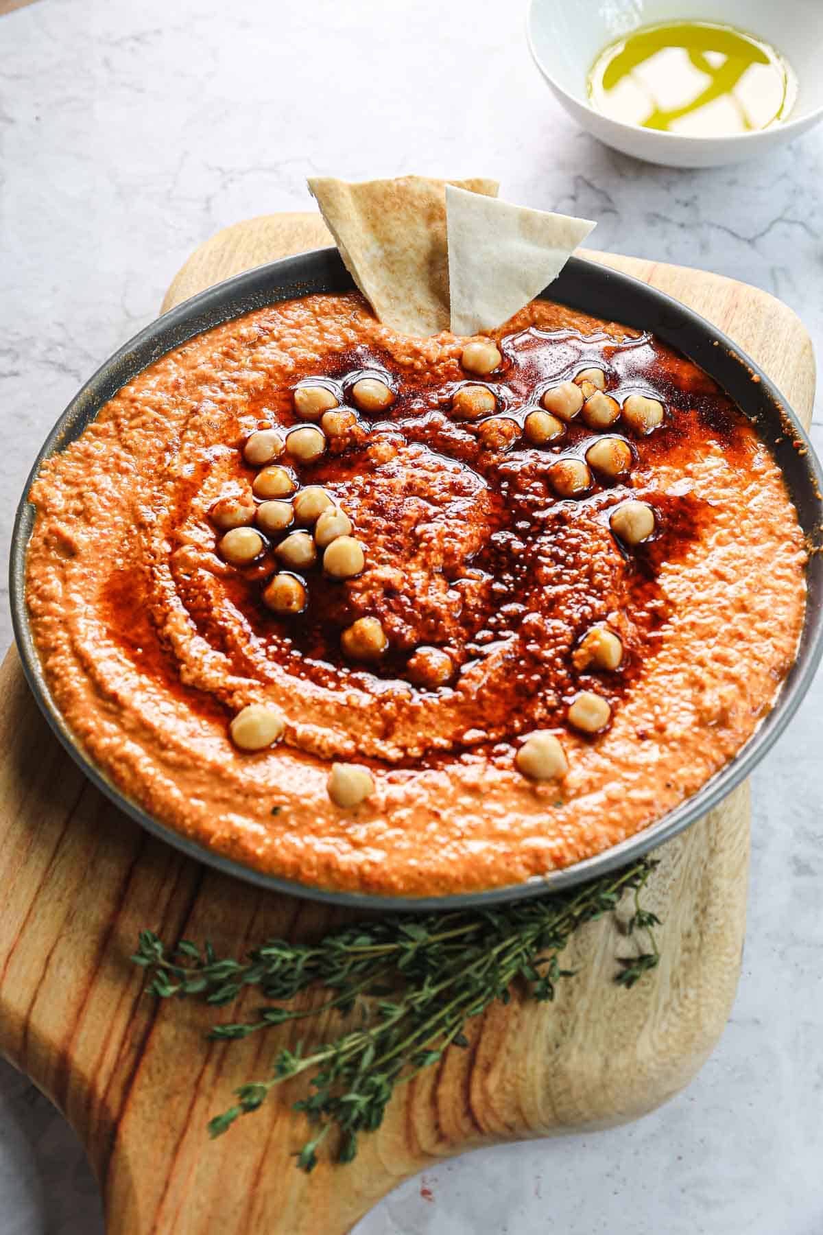 A round dish filled with roasted red pepper hummus, topped with whole chickpeas and a drizzle of oil and spices. Two pieces of pita bread rest on the edge. A small bowl of oil is in the background on a wooden surface, surrounded by sprigs of herbs.