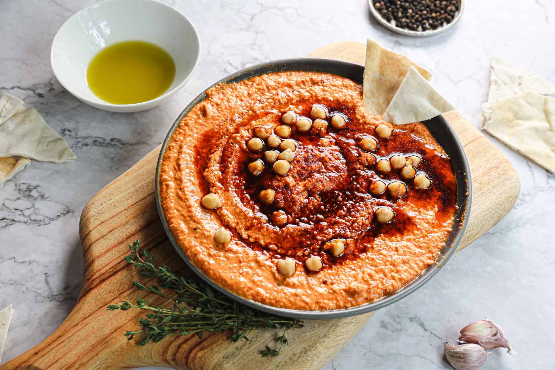 A bowl of roasted red pepper hummus topped with whole chickpeas and spices sits on a wooden board. Alongside are pita wedges, sprigs of thyme, a small bowl of olive oil, garlic cloves, and a dish of peppercorns, all set against a marble surface.