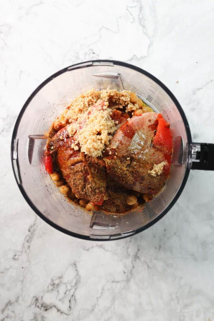 A blender container on a marble countertop, filled with raw chicken pieces, chickpeas, roasted red pepper hummus spices, and crushed garlic.