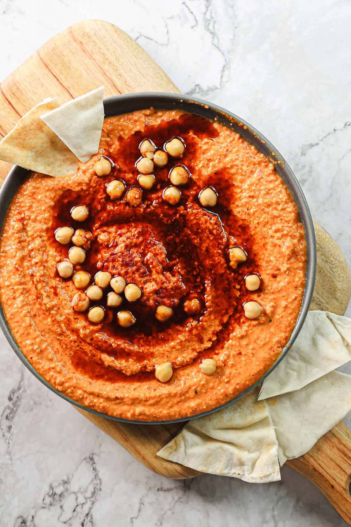 A bowl of roasted red pepper hummus topped with whole chickpeas and drizzled with olive oil sits on a wooden cutting board. Triangular pieces of pita bread are arranged around the bowl, set against a marble surface.