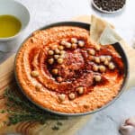 A bowl of creamy roasted red pepper hummus topped with chickpeas and a drizzle of red oil, garnished with bread pieces. The bowl sits on a wooden board with a sprig of thyme. A small bowl of olive oil and peppercorns linger in the background.