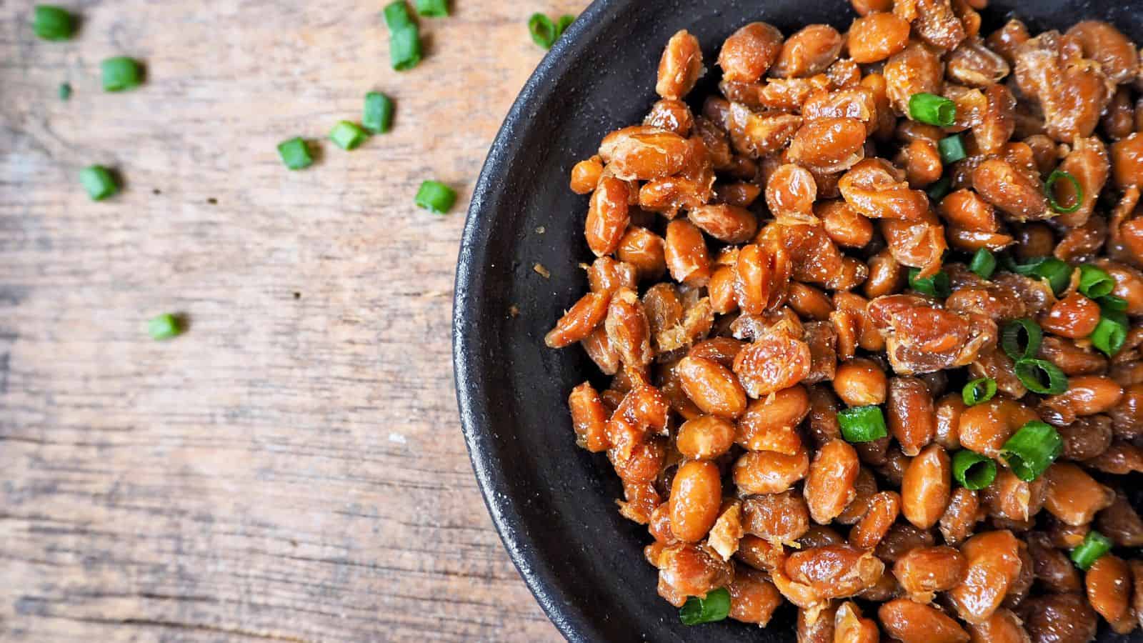 A black bowl filled with savory glazed peanuts topped with chopped green onions. The bowl is placed on a wooden surface, with a few scattered green onion pieces resting beside it.