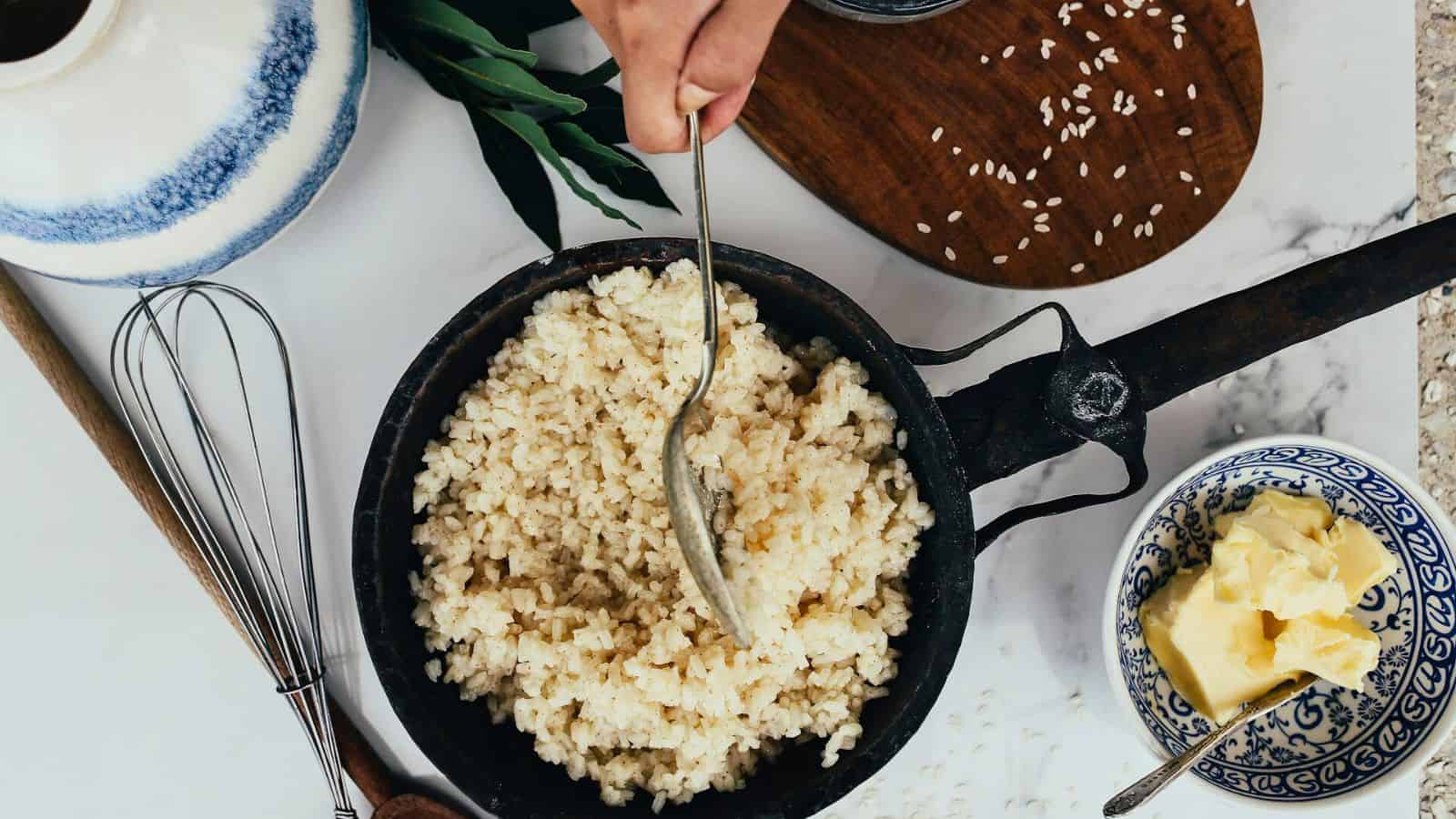 A hand is stirring cooked rice in a black skillet with a metal spoon. Surrounding the skillet are a small bowl with butter, a wooden board with scattered grains of rice, a whisk, and a ceramic jug. Some green leaves are also visible.