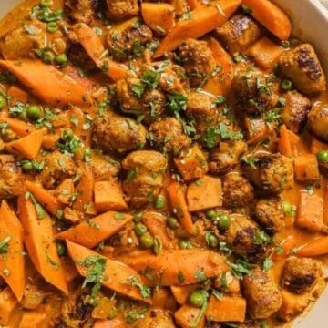 A white skillet filled with a dish of sliced carrots, green peas, and browned meat pieces in a creamy orange sauce evokes taste memories from time-tested recipes. Garnished with chopped herbs adding a touch of green, it transports you to decades ago. A small bowl with herbs sits beside the skillet.