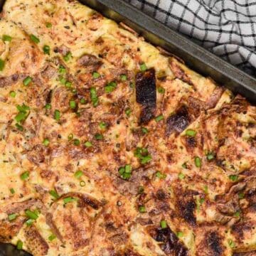 A rectangular metal baking dish contains a golden-brown egg frittata with visible slices of onion and sprinkled with chopped green herbs. A black-and-white checkered cloth is partially visible at the top right corner.