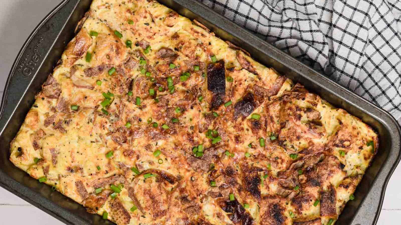 A baked egg and bread casserole in a rectangular metal pan, garnished with chopped green onions. A black and white checkered cloth is partially visible in the background.