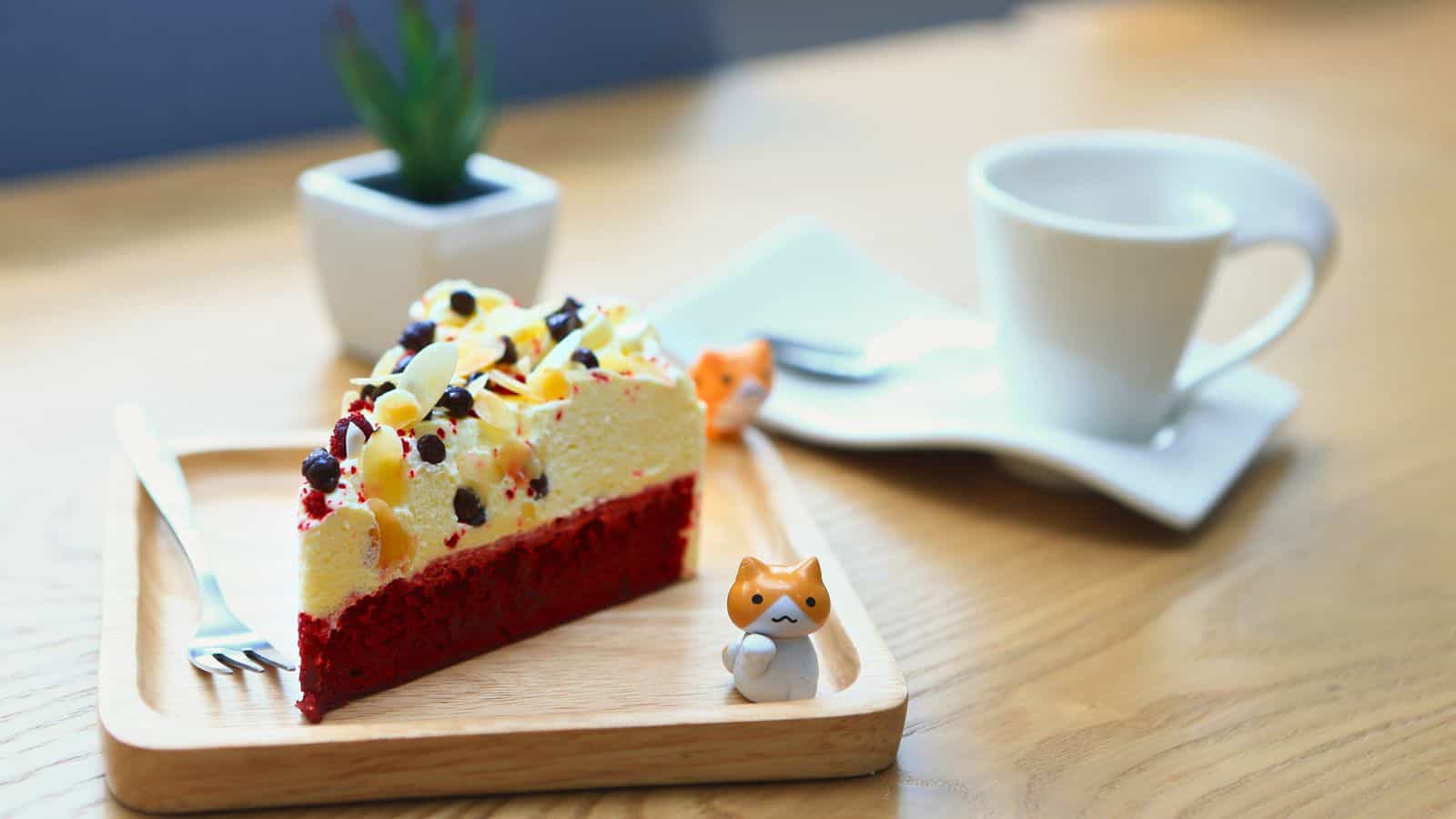 A slice of red velvet cake with a cream topping and assorted fruits is on a wooden plate with a fork. A small ceramic cat figure is beside it. A white cup and saucer are in the background next to a small potted plant.
