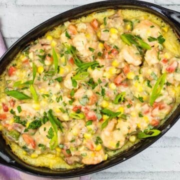 Oval black dish with shrimp casserole on a white brick surface. The casserole contains shrimp, corn, diced tomatoes, and green onions, topped with herbs. A multicolored cloth is partially visible to the side.