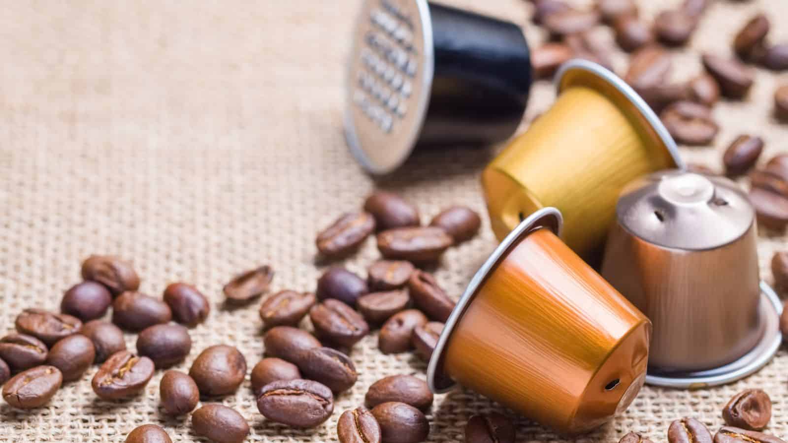 Four espresso coffee capsules in black, gold, copper, and silver rest on a burlap surface surrounded by scattered coffee beans.