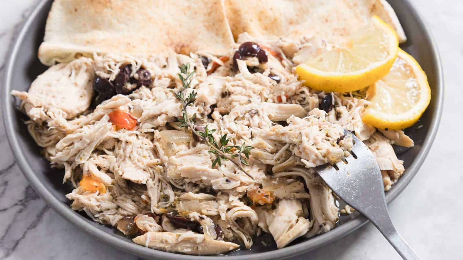 A plate of shredded chicken is garnished with herbs, black olives, and sliced cherry tomatoes. Two lemon wedges and pieces of flatbread accompany the dish. A fork is resting on the edge of the plate.