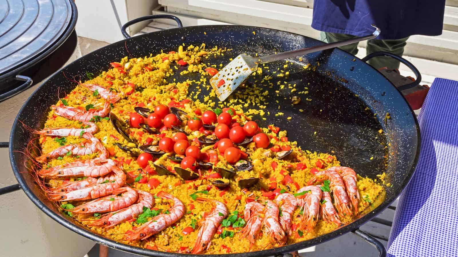 A large pan of vibrant paella featuring saffron-colored rice, arranged with whole shrimp, mussels, and garnished with cherry tomatoes and greenery. A spatula rests in the pan, which sits on a checkered cloth-covered surface.