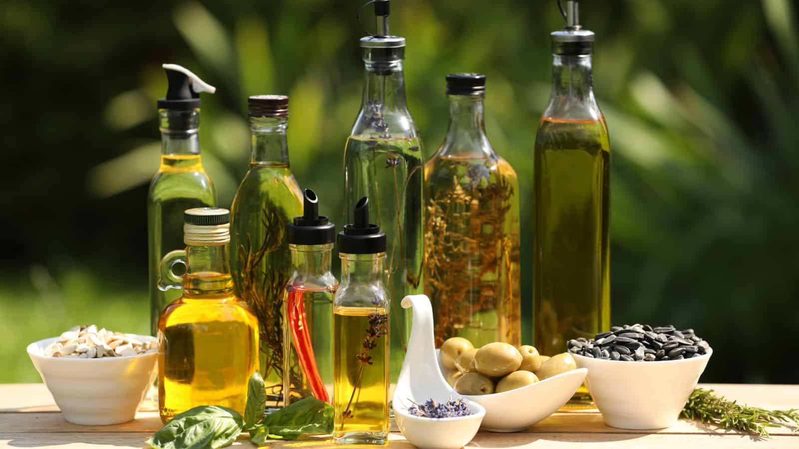 Various bottles of oil with herbs and spices are arranged on a table outdoors. Around them are small bowls containing seeds, nuts, olives, and dried herbs. The background is a blurry, green, natural setting.