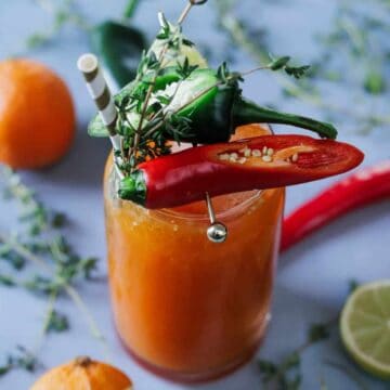 A glass of orange cocktail garnished with red and green chili peppers, herbs, and a straw. Surrounded by whole oranges, a lime half, and scattered thyme sprigs on a light blue background.