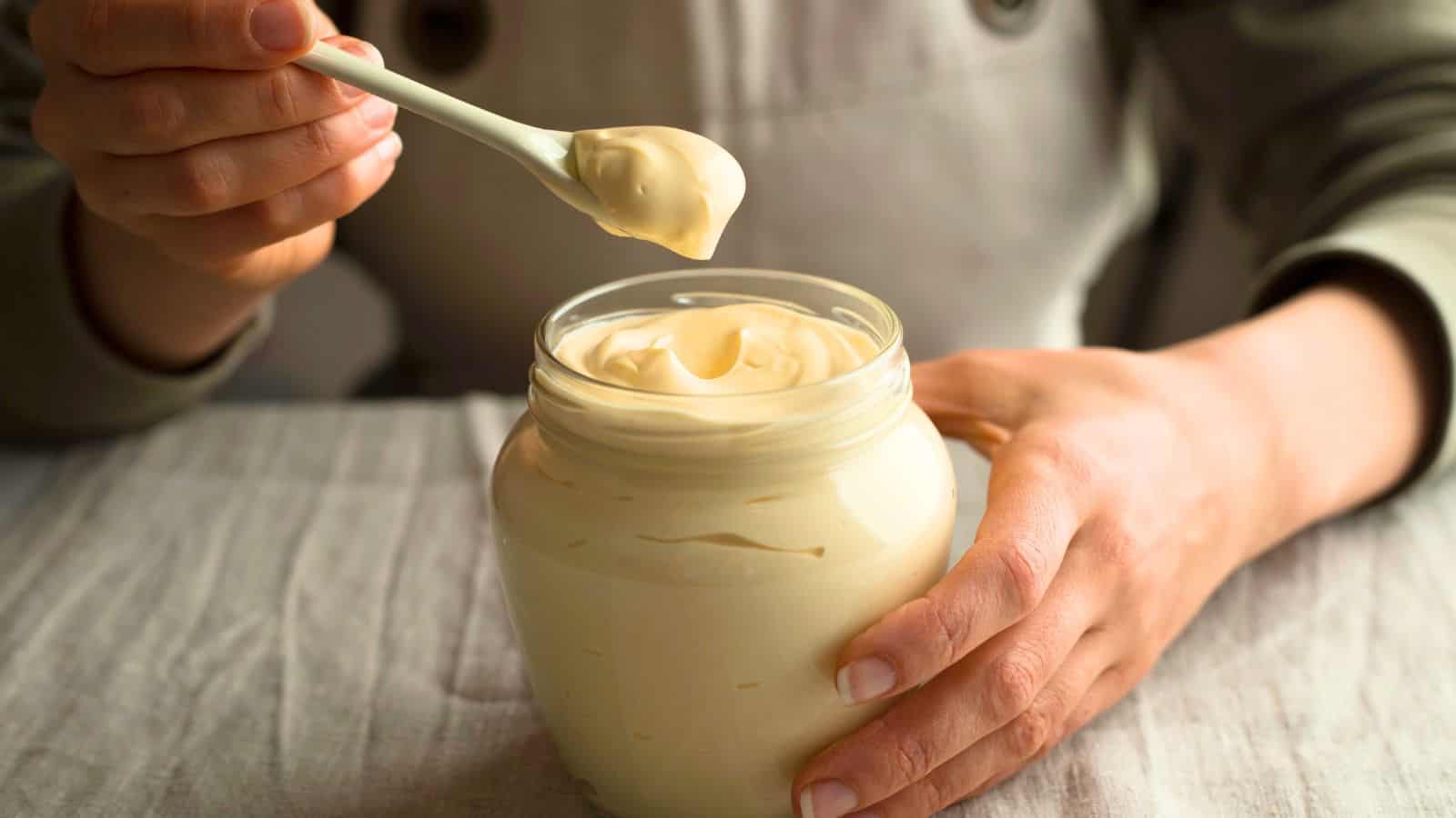 A person holds a jar of mayonnaise on a table, using a spoon to scoop some out. The jar is open, revealing the creamy contents. The person is wearing a gray long-sleeved top, and the table is covered with a light-colored cloth.