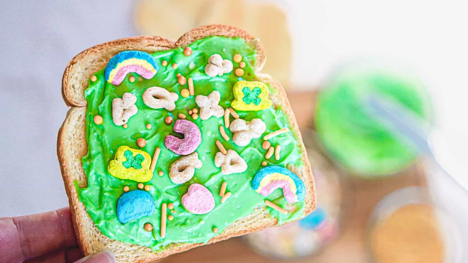 A slice of toast spread with bright green frosting is topped with colorful marshmallow cereal pieces and small sprinkles. A jar of green frosting, a bowl of cereal, and a knife are blurred in the background.