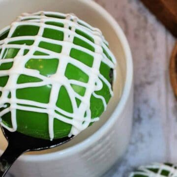 A green round dessert with white drizzle sits on a spoon over a white mug. The mug is on a marble surface with a round wooden board nearby.