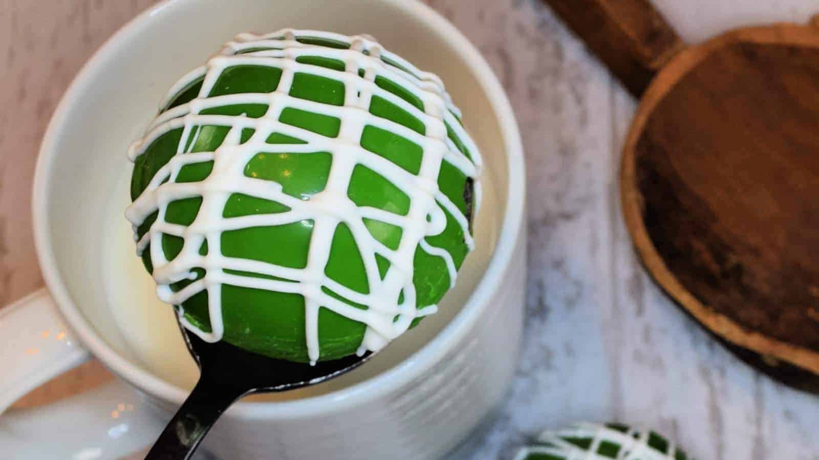 A green round dessert with white drizzle sits on a spoon over a white mug. The mug is on a marble surface with a round wooden board nearby.