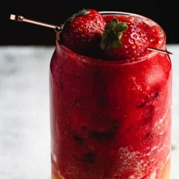 A glass filled with a layered frozen drink, featuring a red slushy texture on top and an orange layer at the bottom. Two whole strawberries garnish the top, placed on a skewer. The background is softly blurred.