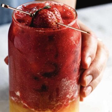 A hand holds a glass containing a layered drink. The top layer is a thick, red slush with a whole strawberry and a garnish, while the bottom is a yellow liquid. A metal straw is inserted into the drink. The surface is light-colored.
