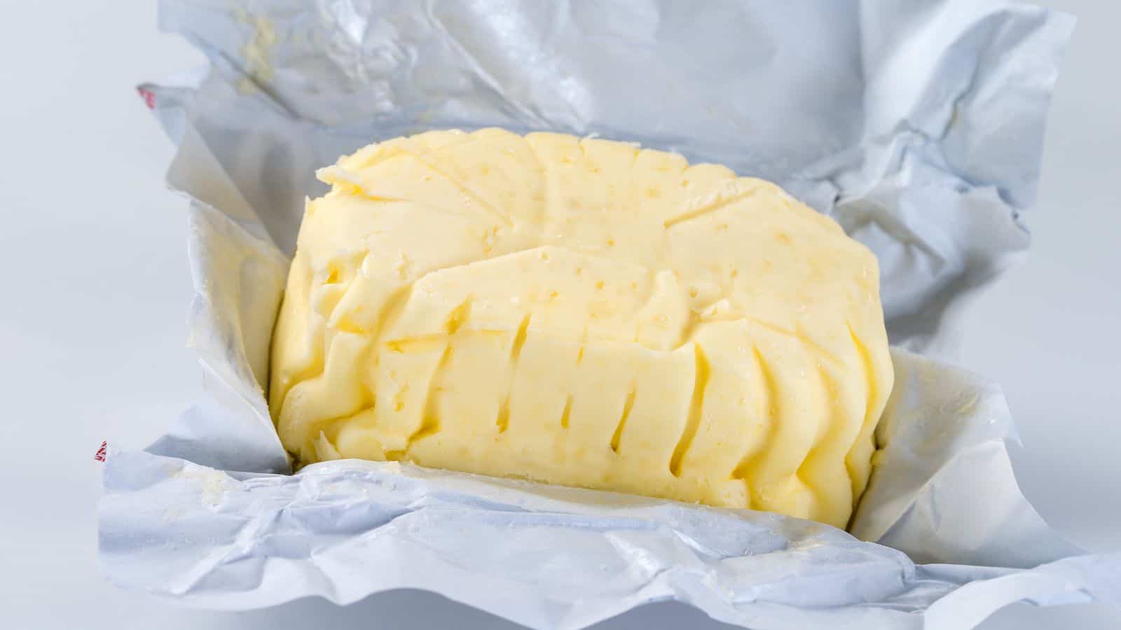 A block of butter is partially unwrapped on gray parchment paper. The butter displays a textured surface with several indentations and small holes. The background is plain and neutral, emphasizing the butter and its wrapping.