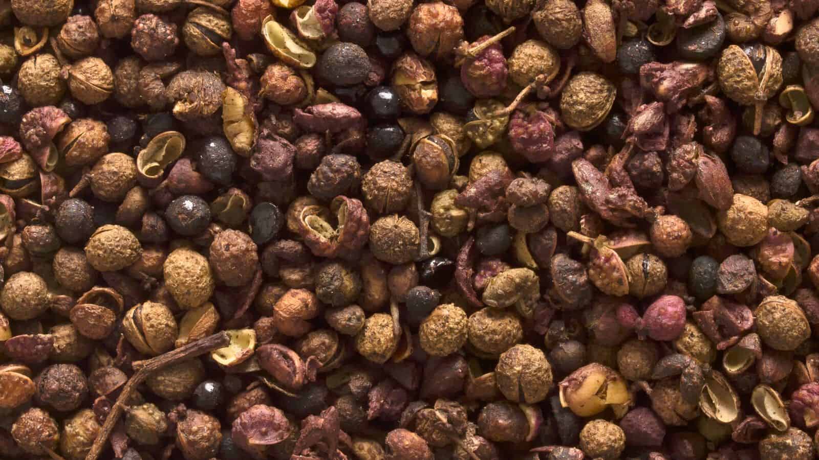 Close-up of a mixture of Sichuan peppercorns, displaying various shades of brown, black, and reddish hues. The peppercorns are round, with textured surfaces, and some split open, revealing inner seeds. A few twig-like stems are visible among them.
