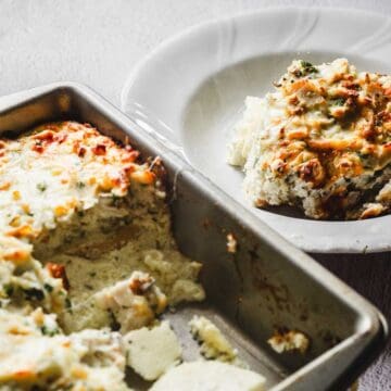 A rectangular baking dish with a baked, cheesy casserole. A serving has been placed on a white plate next to it. The casserole is topped with melted cheese and chopped herbs, with visible layers beneath.