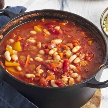 A black pot filled with a tomato-based stew containing white beans, bell peppers, and other vegetables. The pot rests on a dark cloth with a silver spoon nearby. A bowl of couscous sits beside the pot on a white surface.