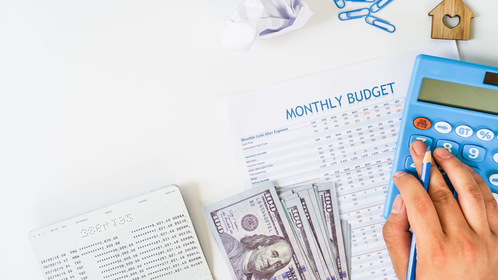 A person uses a blue calculator on a table with a monthly budget sheet, scattered U.S. hundred-dollar bills, a bank statement, blue paperclips, a crumpled paper ball, and a small wooden house figure.