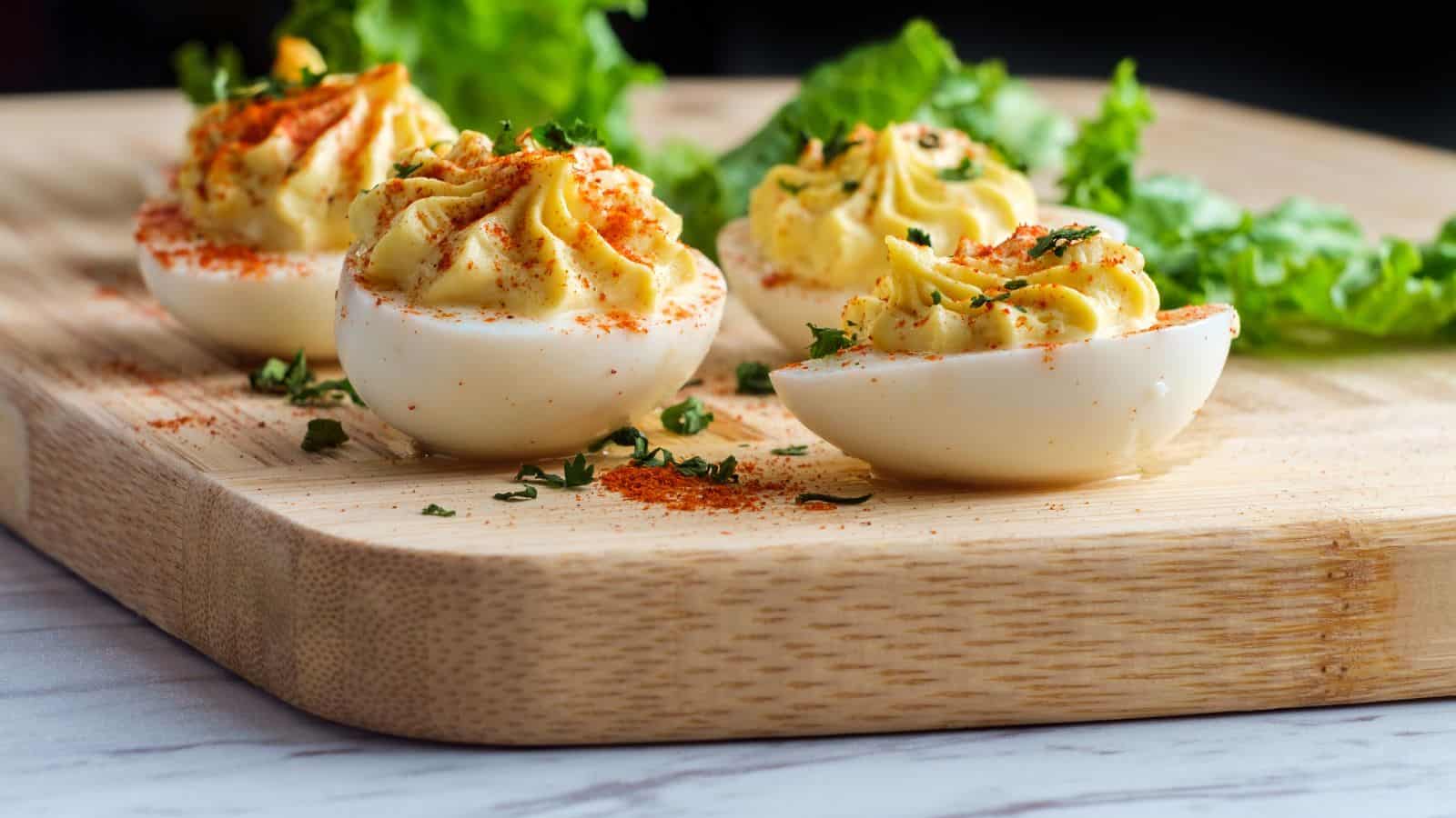 Four deviled eggs, halved and filled with a creamy yellow mixture, are arranged on a wooden cutting board. They are garnished with paprika and chopped herbs. In the background, there are green lettuce leaves.