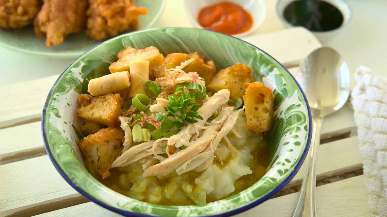 A bowl of porridge topped with shredded chicken, fried tofu pieces, chopped green onions, and crispy crackers on a green-rimmed plate. A spoon is next to the bowl. In the background, there are small bowls with sauces and fried snacks.