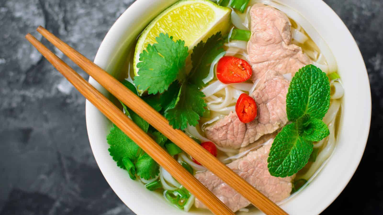 A bowl of pho with rice noodles, slices of beef, lime wedge, coriander, mint leaves, and red chili slices, garnished with green onions. Chopsticks rest on the bowl, and the dark background contrasts with the vibrant colors of the ingredients.