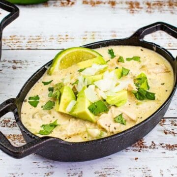 Two black oval dishes filled with a creamy chicken chili stew, topped with chopped cilantro, avocado, onion, and a lime wedge, placed on a white, weathered wooden surface. A white and gray striped cloth is partially visible in the lower left corner.