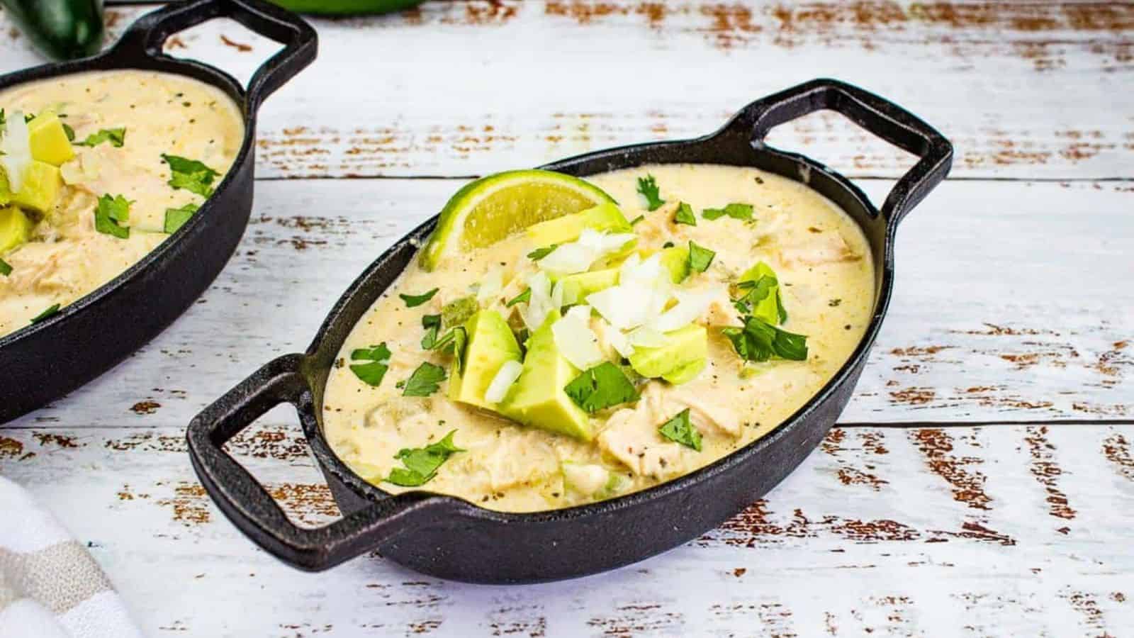Two black oval dishes filled with a creamy chicken chili stew, topped with chopped cilantro, avocado, onion, and a lime wedge, placed on a white, weathered wooden surface. A white and gray striped cloth is partially visible in the lower left corner.