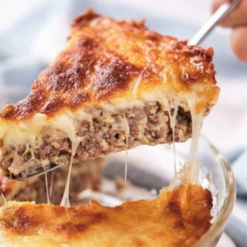 A slice of moussaka being lifted from a glass baking dish. The slice shows layers of ground meat and a golden-brown cheese topping, with melted cheese stretching as it's lifted. A hand holds the server.