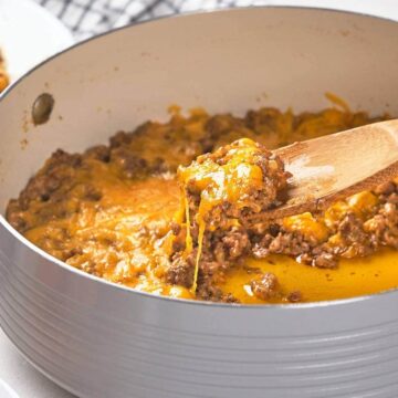 A saucepan filled with a mixture of ground meat and melted cheese is placed on a countertop. A wooden spatula is lifting a portion from the pan. In the background, a white towel with black stripes and a partial view of a white plate can be seen.