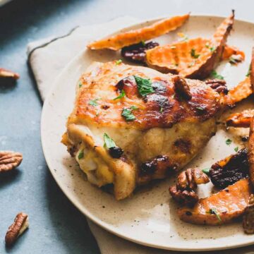 A plate with roasted chicken thigh, seasoned sweet potato wedges, and pecans exemplifies easy sheet pan meals. The dish is garnished with chopped herbs, while a small bowl of pecans sits nearby on the gray surface.