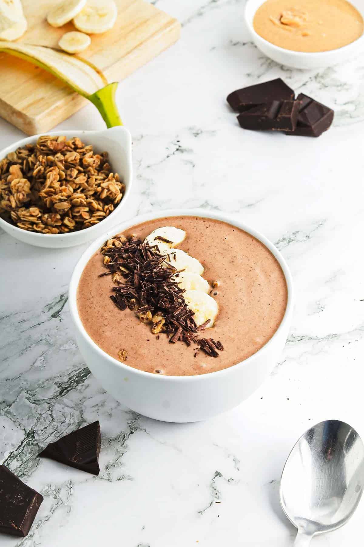 A chocolate peanut butter smoothie bowl sits on a marble surface, topped with sliced bananas and chocolate shavings. Nearby are granola, chocolate pieces, a spoon, and a small bowl of peanut butter. A wooden board holds additional banana slices.