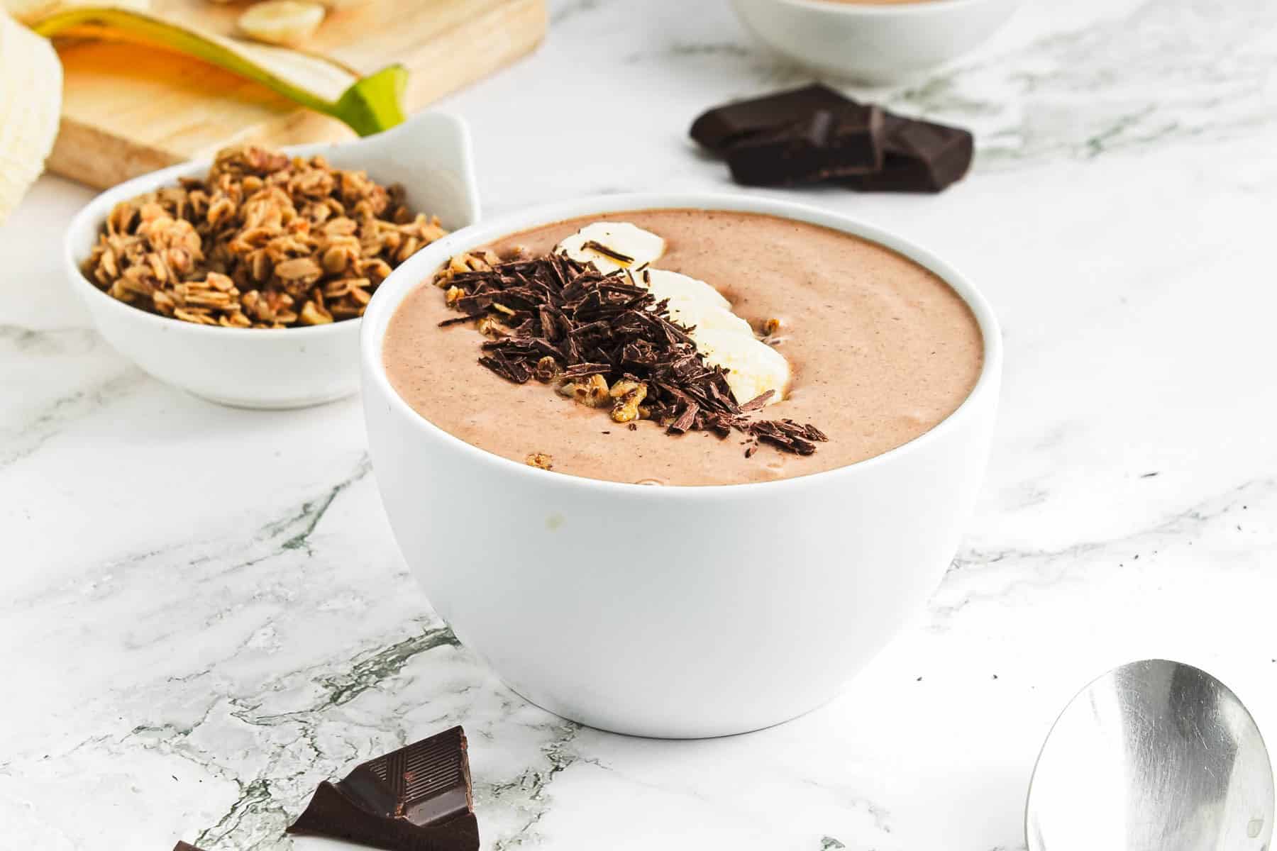 A white bowl filled with a creamy chocolate peanut butter smoothie topped with banana slices and shaved chocolate, set on a marble surface. A small bowl of granola and dark chocolate pieces are in the background. A spoon lies nearby.