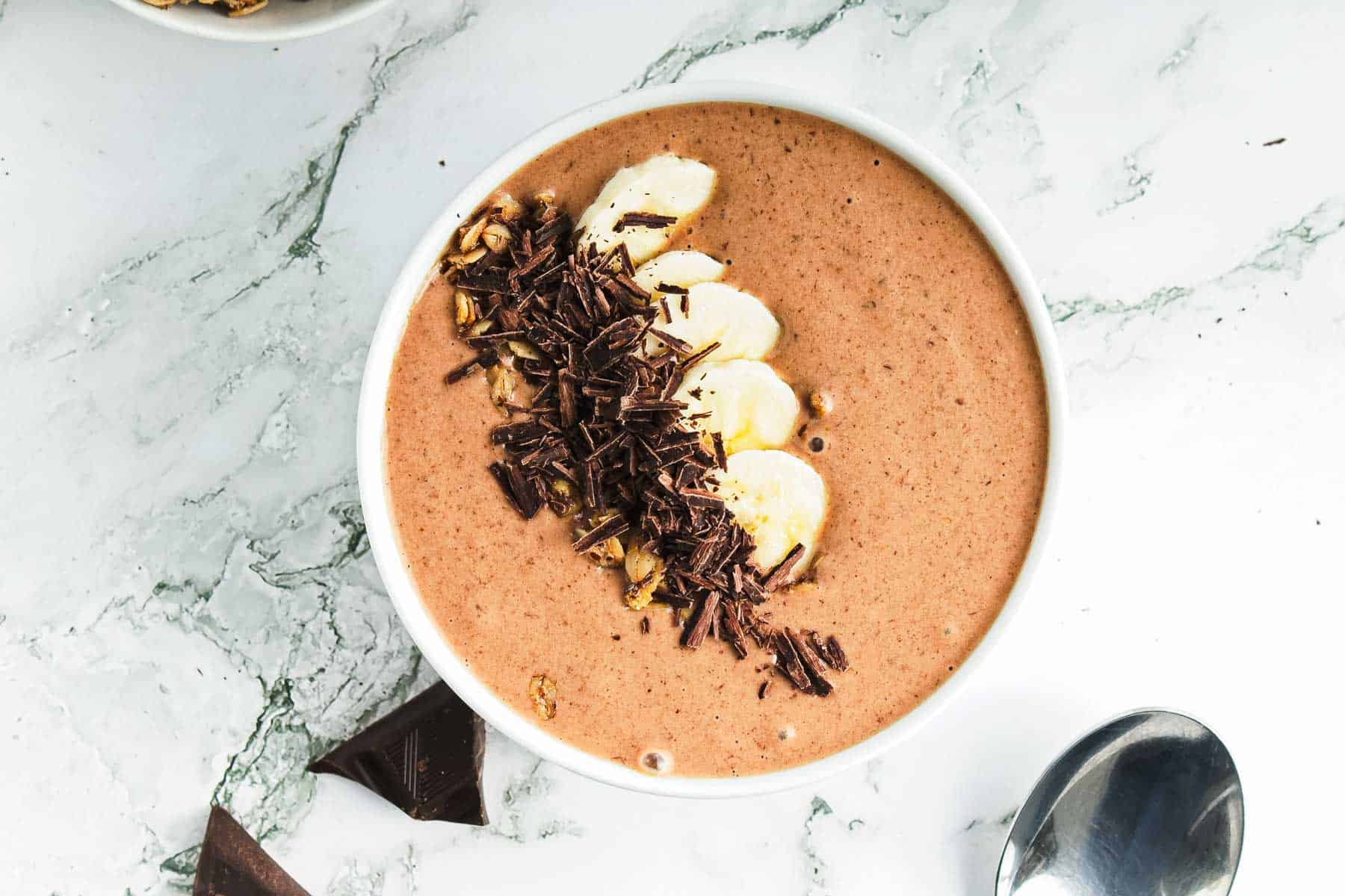 A bowl of chocolate peanut butter smoothie topped with sliced bananas, shredded chocolate, and granola rests on a marble surface. A spoon is nearby.