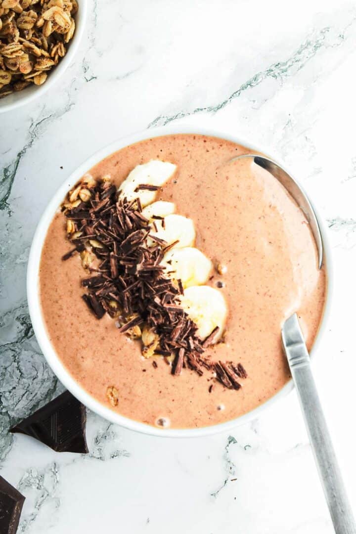 A bowl filled with a chocolate peanut butter smoothie topped with banana slices, granola, and chocolate shavings. A spoon rests in the bowl, inviting a taste. A small dish of extra granola is in the top left corner on a white marble surface.