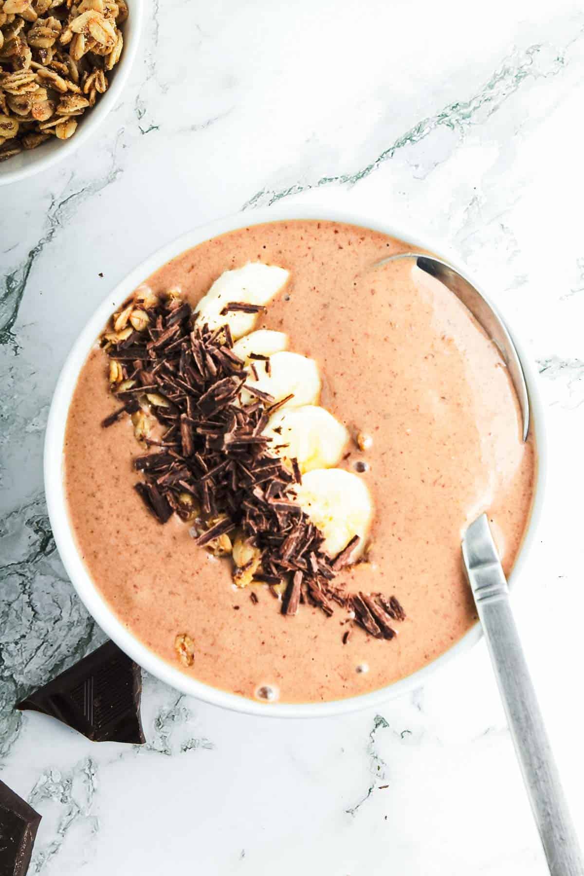 A bowl filled with a chocolate peanut butter smoothie topped with banana slices, granola, and chocolate shavings. A spoon rests in the bowl, inviting a taste. A small dish of extra granola is in the top left corner on a white marble surface.