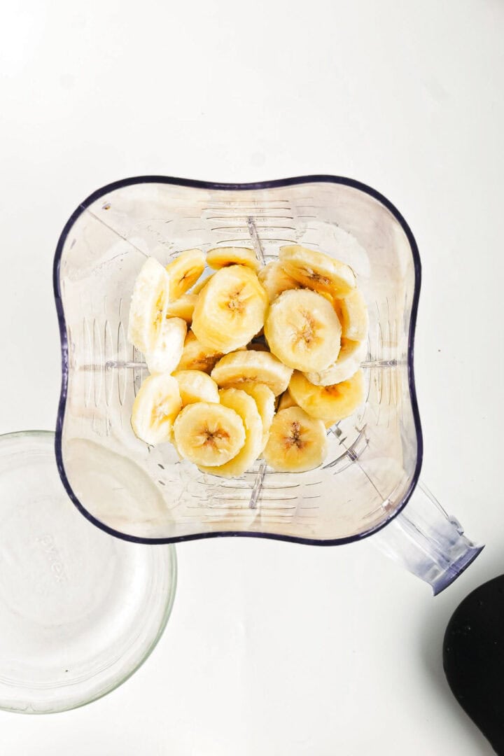 Sliced bananas are placed at the bottom of a blender jug on a white surface, ready to blend into a delicious Chocolate Peanut Butter Smoothie. The jug's clear walls showcase the ingredients, while an empty glass bowl waits beside it for serving.