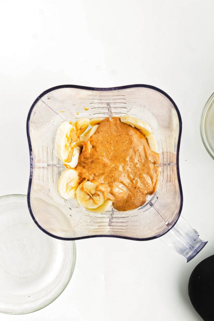 A blender containing sliced bananas and creamy peanut butter—perfect for crafting a Chocolate Peanut Butter Smoothie Bowl. The bananas are mostly on the left, while the peanut butter is centered. Nearby, an empty glass bowl awaits its delicious transformation.