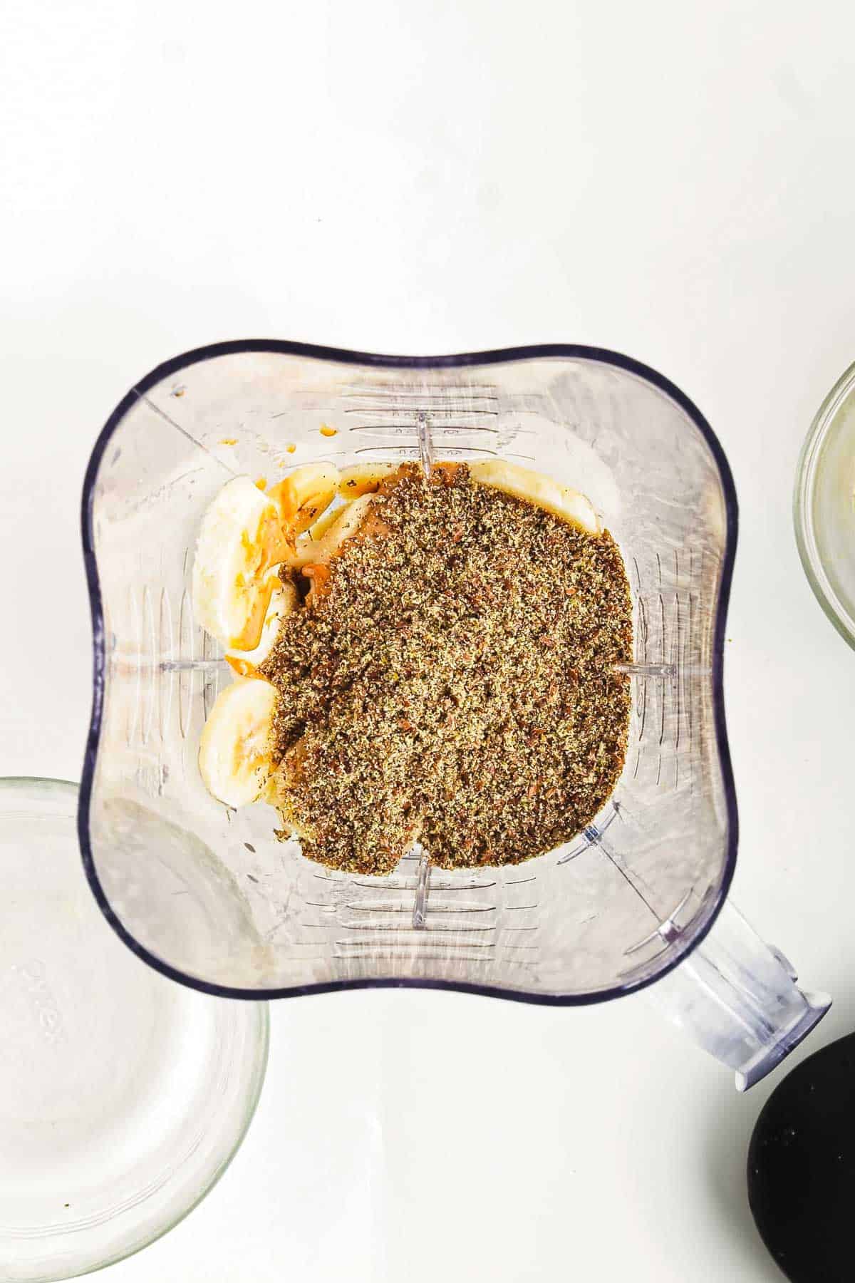 A blender viewed from above, containing banana slices, peanut butter, and a pile of ground flaxseed—ingredients destined for a delicious Chocolate Peanut Butter Smoothie Bowl. The blender rests on a white surface, with parts of two empty glass bowls visible nearby.