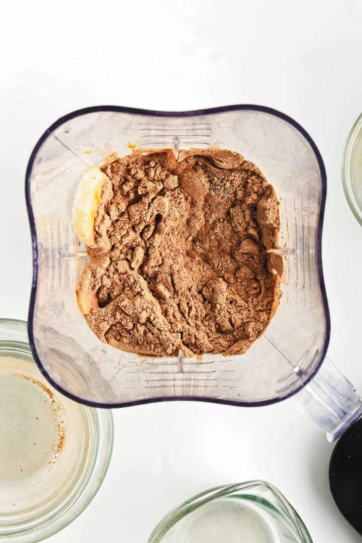 Top view of a blender container filled with brown powder, possibly chocolate or cocoa, on a white surface. The setup hints at the beginnings of a tempting chocolate peanut butter smoothie bowl, with empty glass bowls eagerly awaiting their fill.
