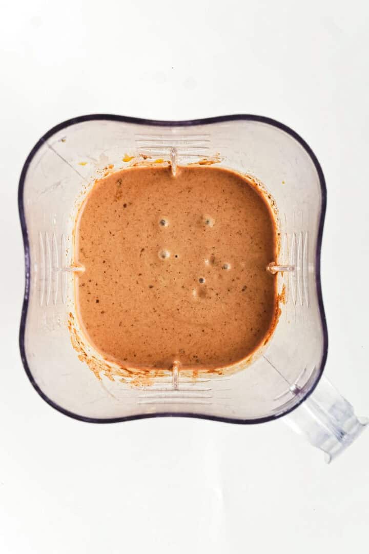 A blender jar holds a thick, brownish-orange mixture reminiscent of a chocolate peanut butter smoothie bowl, with a few air bubbles visible on the surface. The background is plain white.