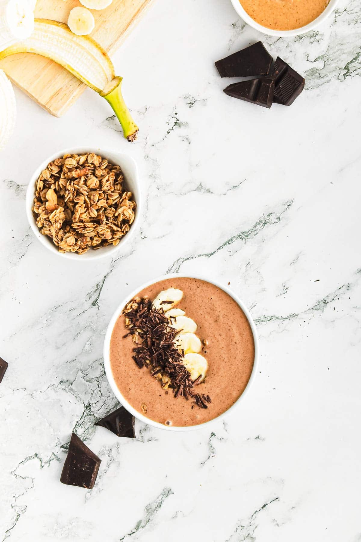 A Chocolate Peanut Butter Smoothie Bowl with sliced bananas and chocolate shavings sits on a marble surface. Nearby, a bowl of granola and chunks of dark chocolate await, while a partially peeled banana rests on a wooden board in the upper left corner.