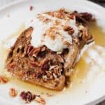 A slice of gluten-free French toast casserole topped with whipped cream, chopped pecans, and sprinkled cinnamon on a white plate. Syrup is drizzled over the toast, pooling around it.